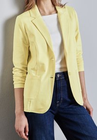 Gele katoenen blazer met een enkele knoop, gespleten revers, twee voorzakken en lange mouwen, gecombineerd met een wit topje en donkere jeans.