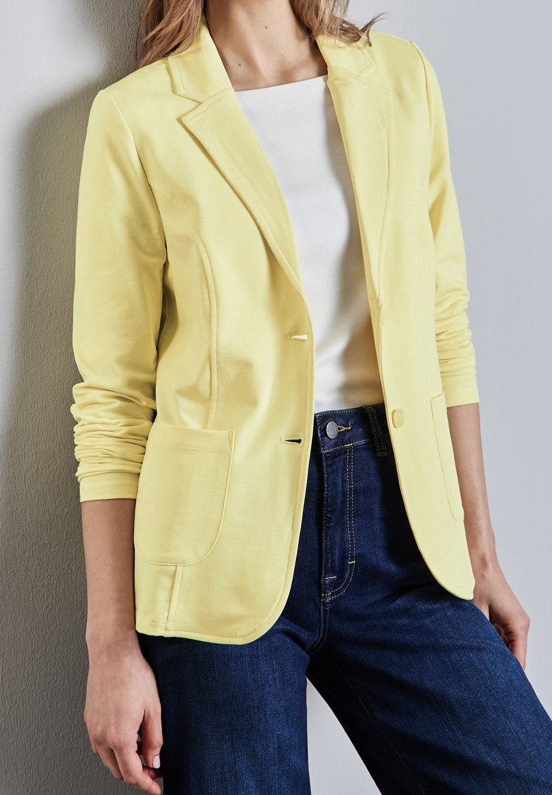 Gele katoenen blazer met een enkele knoop, gespleten revers, twee voorzakken en lange mouwen, gecombineerd met een wit topje en donkere jeans.