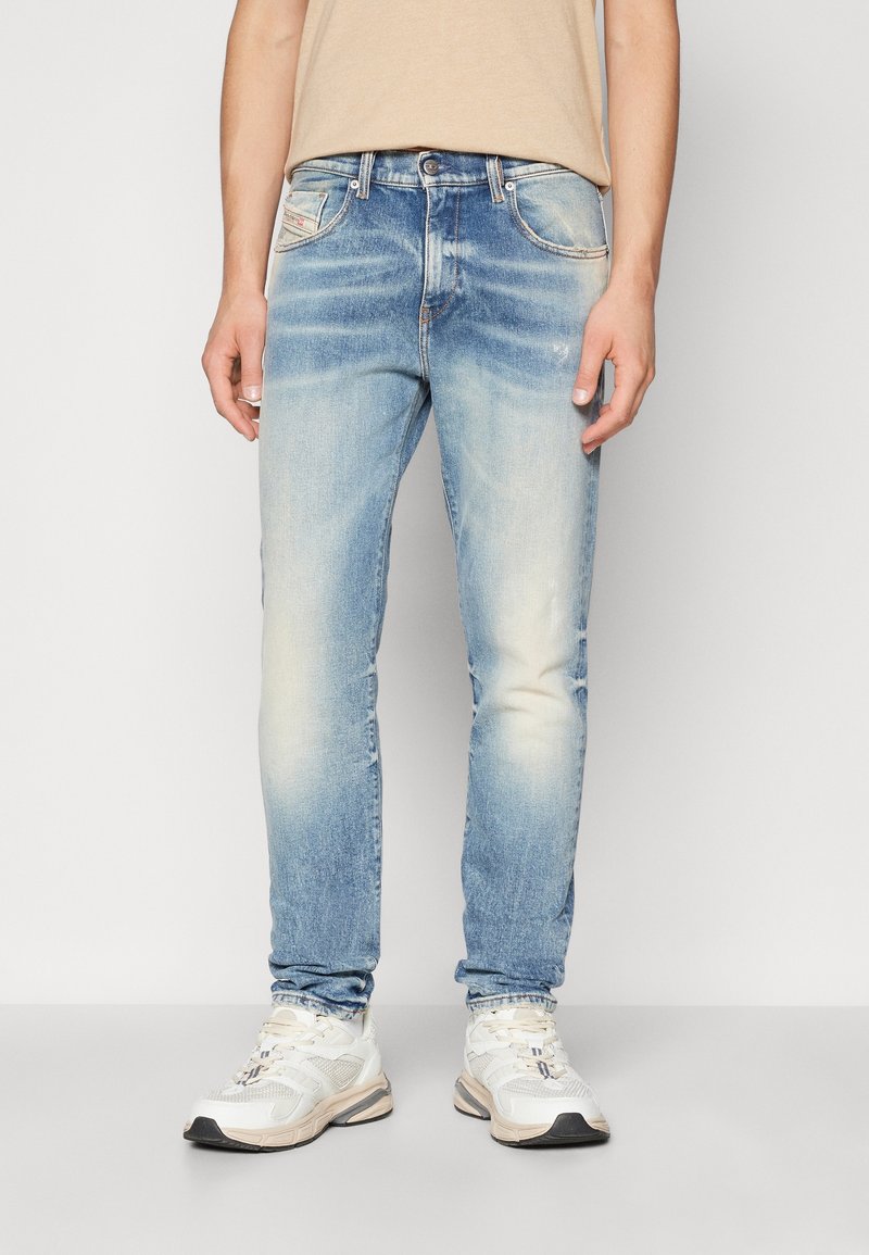 Homme portant un jean bleu clair délavé, un t-shirt beige et des baskets blanches, debout devant un fond clair uni.