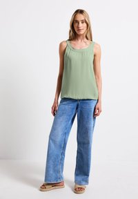 Světlé zelené bezrukávové tričko se nařaseným výstřihem, zkombinované s volnými světlými modrými džíny a pletenými béžovými sandály.