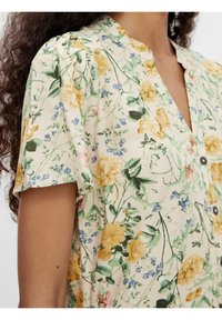 Blouse à manches courtes florale de couleur crème, ornée de fleurs jaunes et bleues, avec un col et des boutons noirs pour la fermeture. Tissu à la texture douce.