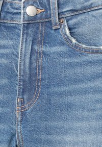 Gros plan sur l’avant d’un jean en denim bleu avec bouton en métal, passants de ceinture, coutures et bord effiloché sur la poche avant droite.