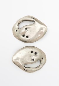 Metallisch zilveren gespen met organische vormen, met een glanzend oppervlak, onregelmatige uitsparingen en kleine gaatjes voor textuur en detail.