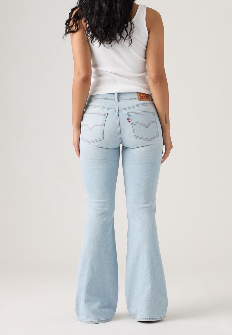 Femme aux longs cheveux bouclés portant un débardeur blanc et un jean évasé bleu clair, debout de dos sur un fond uni.