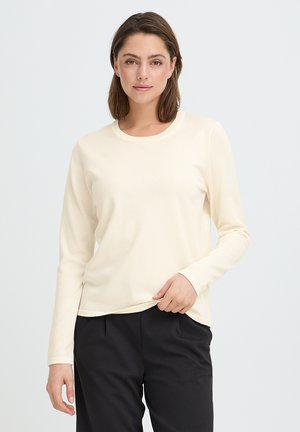 Young woman with shoulder-length brown hair wearing a cream long-sleeve top and black pants, standing against a plain light background.