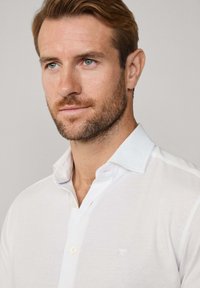 Biała koszula z jasnoniebieskim kołnierzykiem, z pojedynczym zapięciem na guzik i małym haftem po lewej stronie. Gładka faktura materiału.