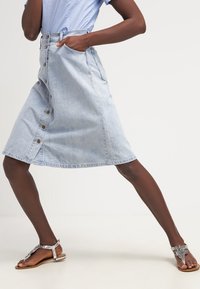Helleblaue Denim-A-Linien-Rock mit Knopfleiste, zwei Seitentaschen und einem Saum, der bis zur Mitte der Wade reicht. Strukturierter Stoff und lässiges Design.