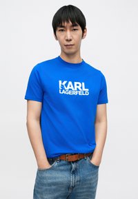 Zils īss piedurkņu t-krekls ar baltu “KARL LAGERFELD” logotipu priekšpusē. Apaļš kakls, izgatavots no gluda kokvilnas materiāla.
