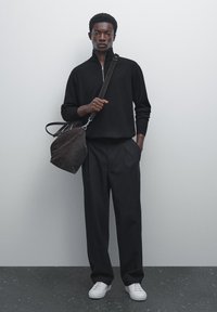 Black zip-up sweater, tailored black trousers, white sneakers, and a dark brown shoulder bag with a textured finish. Plain background.