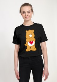 Camiseta negra con un oso de dibujos animados que tiene un corazón en el vientre. El oso es marrón con una cara blanca y mejillas sonrosadas.