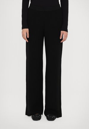 Persoon draagt zwarte, wijde gebreide broek en zwarte schoenen met studs, staand tegen een effen witte achtergrond.