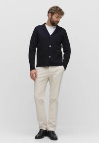 Marineblauwe gebreide cardigan met een sjalkraag, voorzien van voorzakken en witte knopen, gecombineerd met crèmekleurige broeken en zwarte loafers.