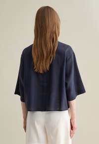 Navy blue linen cropped jacket with short sleeves, featuring a subtle textured pattern on the sides, paired with light-colored, loose-fitting pants.