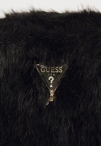 Fausse fourrure noire avec de longues fibres ; présente une étiquette en triangle en or et noir avec "GUESS U.S.A." et un design de point d'interrogation.
