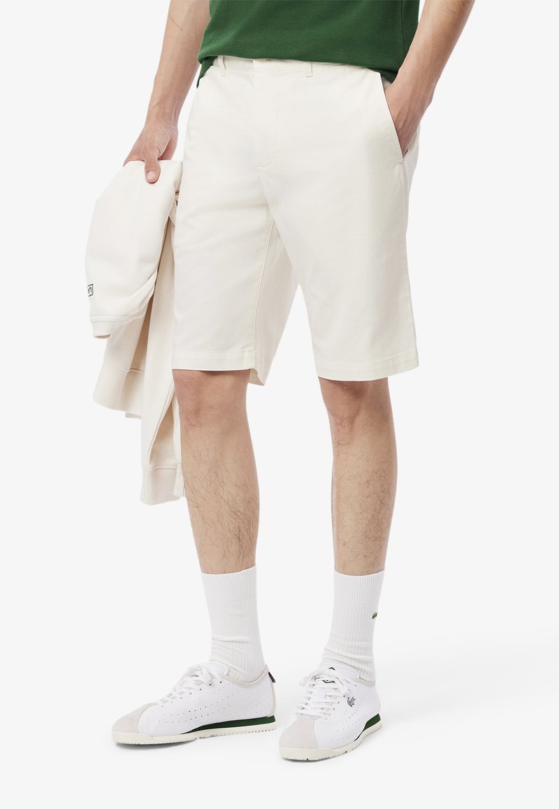 Pantaloni corti crema da uomo con una parte anteriore piatta, tasche laterali e una texture liscia, abbinati a una maglietta verde e sneaker bianche. Tono chiaro.