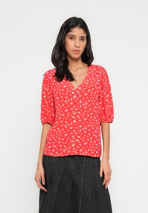 Κόκκινη μπλούζα με λουλουδάτο σχέδιο, κοντά φουσκωτά μανίκια, V-λαιμόκοψη και κουμπιά στο μπροστινό μέρος. Λουλουδάτο μοτίβο σε μαλακό ύφασμα. Συνδυάζεται με σκοτεινή, πλισέ φούστα.