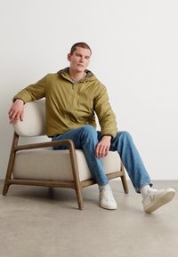 Hellgrüner Kapuzenjacke aus leichtem Material, mit Reißverschluss vorne, kombiniert mit hellblauen Jeans und weißen Sneakern, sitzt auf einem Holzstuhl.