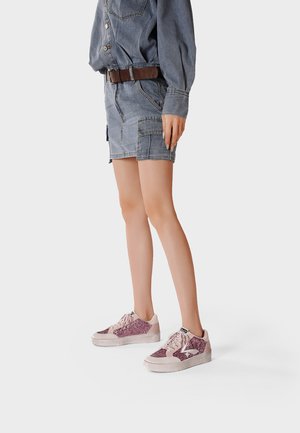 Donna con gonna e giacca di jeans con cintura marrone, in piedi, scarpe da ginnastica glitter rosa con lacci su sfondo uniforme.