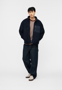 Νέος άνδρας που φοράει μπλε φλις μπουφάν με φερμουάρ στο τσεπάκι του στήθους, ριγέ πουκάμισο, φαρδιά σκούρα τζιν και κρεμ αθλητικά παπούτσια, στέκεται χαλαρός.