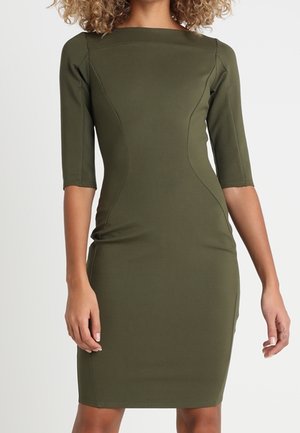 Frau trägt ein eng anliegendes olivgrünes knielanges Kleid mit Ellbogen-langen Ärmeln und Boot-Ausschnitt, steht vor einfachem Hintergrund.