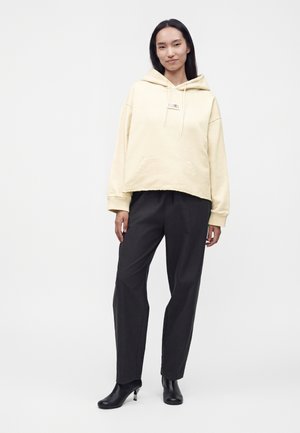 Γυναίκα που φοράει ένα κρεμ oversized φούτερ, μαύρο ίσιο παντελόνι και μαύρα μποτάκια με τακούνι, στέκεται σε ένα απλό ανοιχτό φόντο.