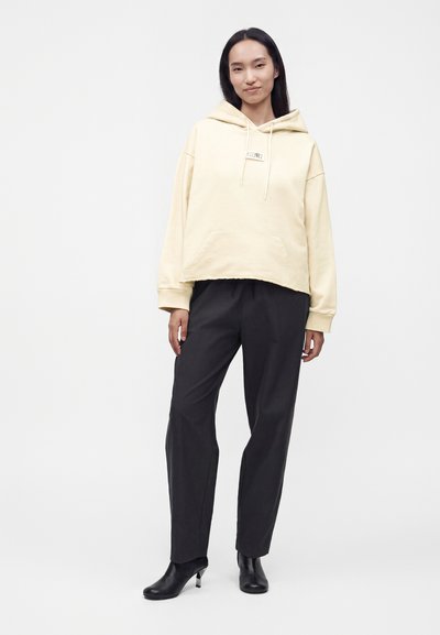 Γυναίκα που φοράει ένα κρεμ oversized φούτερ, μαύρο ίσιο παντελόνι και μαύρα μποτάκια με τακούνι, στέκεται σε ένα απλό ανοιχτό φόντο.