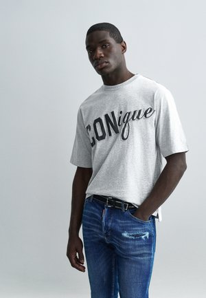 Man draagt een grijs T-shirt met de tekst "CONique" en gescheurde blauwe spijkerbroek, staand met één hand in de zak tegen een effen lichte achtergrond.