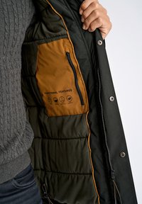 Svart pufferjacka med olivgrön foder, med en brun ficka med dragkedja och funktionella funktionsikoner tryckta på den.