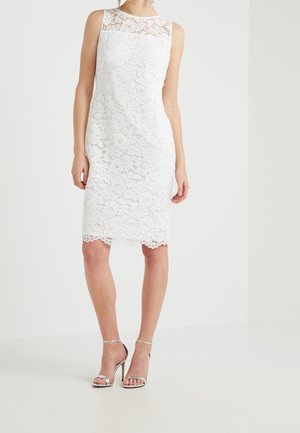 Mouwloze witte knielange kanten jurk met geschulpte zoom, gedragen met zilverkleurige sandalen met hoge hakken, op een effen achtergrond.