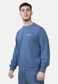 Uomo che indossa una felpa blu e pantaloni abbinati con un piccolo triangolo bianco e un logo a X sul petto, in piedi davanti a uno sfondo semplice.