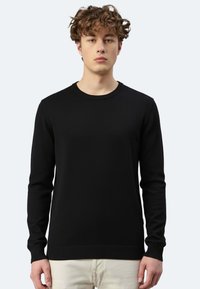Schwarzer Strickpullover mit langen Ärmeln, geripptem Halsausschnitt und Saum. Getragen über hellen Hosen aus einem strukturierten Stoff.