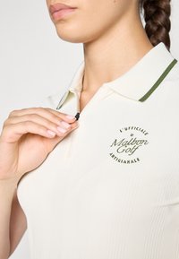 Hvit polo skjorte med ribbestruktur, grønn kragekant og brodert "Malbon Golf"-logo på brystet. Glidelåsdetalj nær halsen.