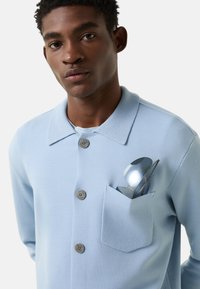 Lyseblå strikket skjorte med knapper, krave og lomme, med grå knapper og solbriller anbragt i brystlommen.