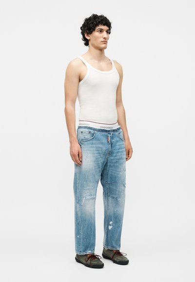 Άνδρας στέκεται φορώντας λευκή φανέλα, ξεθωριασμένο σκισμένο μπλε τζιν, πράσινα παπούτσια, με σγουρά σκούρα μαλλιά σε απλό λευκό φόντο.