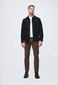 Czarna koszula w kratę z guzikami noszona na białej koszulce, zestawiona z brązowymi, dopasowanymi spodniami i czarnymi, wiązanymi butami. Prosty, nowoczesny design.