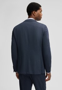 Granatowa marynarka wykonana z fakturowanej tkaniny, o klasycznym kroju z wciętymi klapami i dwoma zapięciami na guziki, przedstawiona od tyłu.