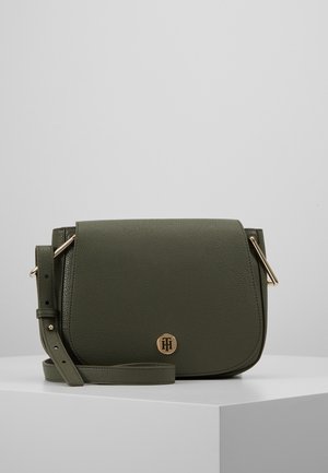 Bolso bandolera de cuero verde oscuro con correa ajustable y emblema de logo en tono dorado en la solapa frontal, exhibido sobre una superficie blanca.