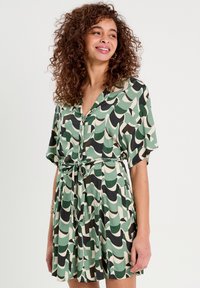 Robe à manches courtes dans un motif de vagues vert, noir et crème. Dotée d'un col à boutons et d'un détail de taille nouée. Tissu léger.