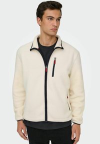 Junger Mann, der eine fleecefarbene Jacke in Creme mit schwarzen und roten Reißverschlussdetails trägt. Er steht entspannt mit Händen und schaut nach rechts.