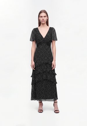Femme aux cheveux raides porte une robe noire à pois avec un col en V, des manches volantes, une jupe à volants superposés et des sandales noires à talons hauts.