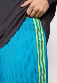 Blaue Sportshorts aus strukturiertem Stoff, mit vertikalen Streifen und gelben Akzenten entlang der Seitennähte.