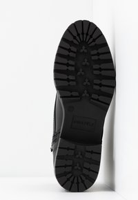 Chaussures noires avec une semelle en caoutchouc texturée ; elles présentent un motif de bande de roulement épais et un détail de marque embossé. Le design est minimaliste et fonctionnel.