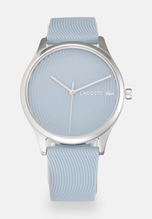 Round Lacoste watch with a textured light blue silicone strap and matching dial, silver-tone hands, and logo, featuring a minimalist design.
