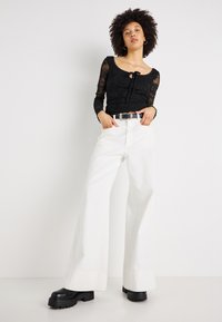 Μαύρη δαντελένια μπλούζα με μακριά μανίκια, λεπτομέρεια με κορδόνι και σφηκοφωλιά λαιμόκοψη, συνδυασμένη με λευκούς παντελόνες υψηλής μέσης και μαύρες πλατφόρμες μποτάκια.