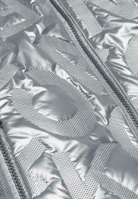 Veste en duvet argentée avec des motifs en relief, texture brillante et une fermeture éclair centrale avec des détails noirs. Sections et surfaces distinctes visibles.