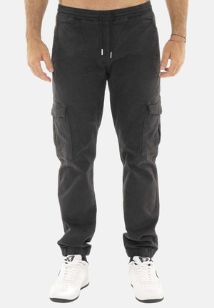 Homme portant un pantalon cargo noir à taille coulissante avec cordon et poignets élastiques, associé à des baskets blanches sur fond blanc.
