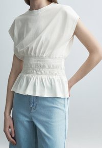 Femme portant un haut blanc sans manches avec taille froncée et un jean en denim bleu clair, debout devant un fond clair uni.