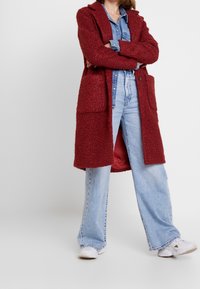 Abrigo largo texturizado rojo con dos bolsillos delanteros, llevado sobre una camisa vaquera y pantalones vaqueros acampanados azules. Unas zapatillas blancas completan el conjunto.