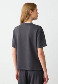Grijze casual t-shirt met korte mouwen, ronde halslijn en subtiele versieringen van kleine studs op de schouders. Een gladde textuur en een relaxte pasvorm.