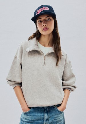 Jeune femme portant un sweat-shirt gris à fermeture éclair quart, un jean bleu et une casquette bleu marine avec le mot "Fearless" brodé en rouge.